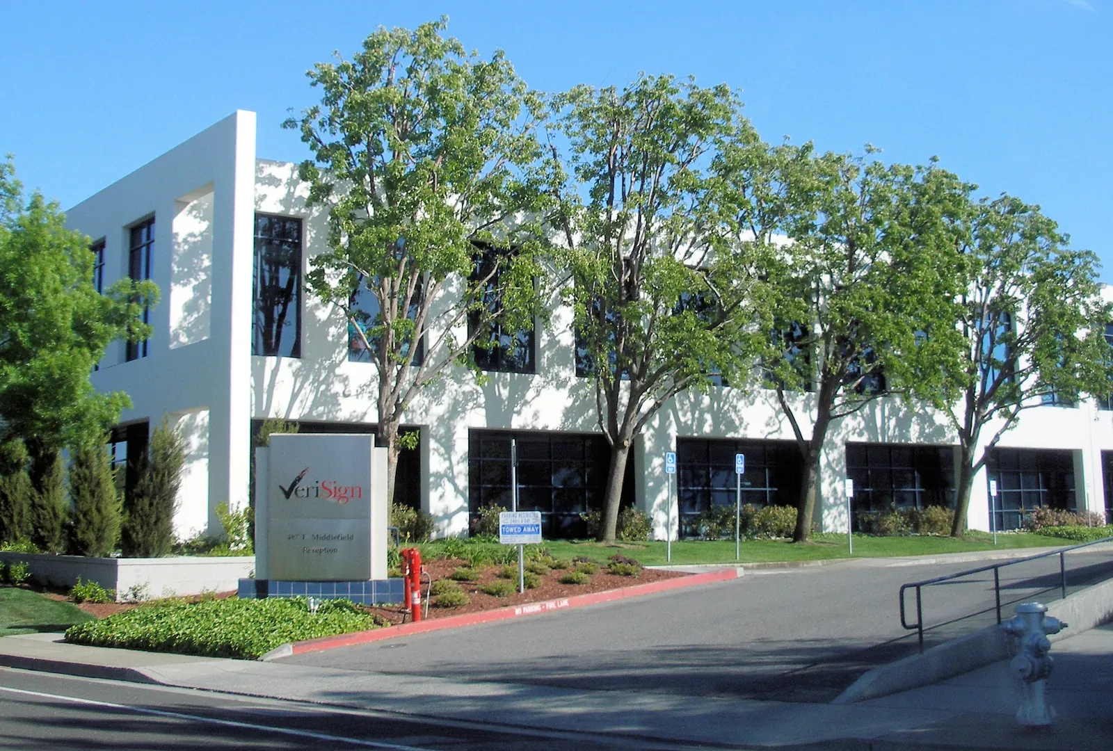 A white two-storey office building behind a row of trees, with a VeriSign company sign on a lawn beside the road in front