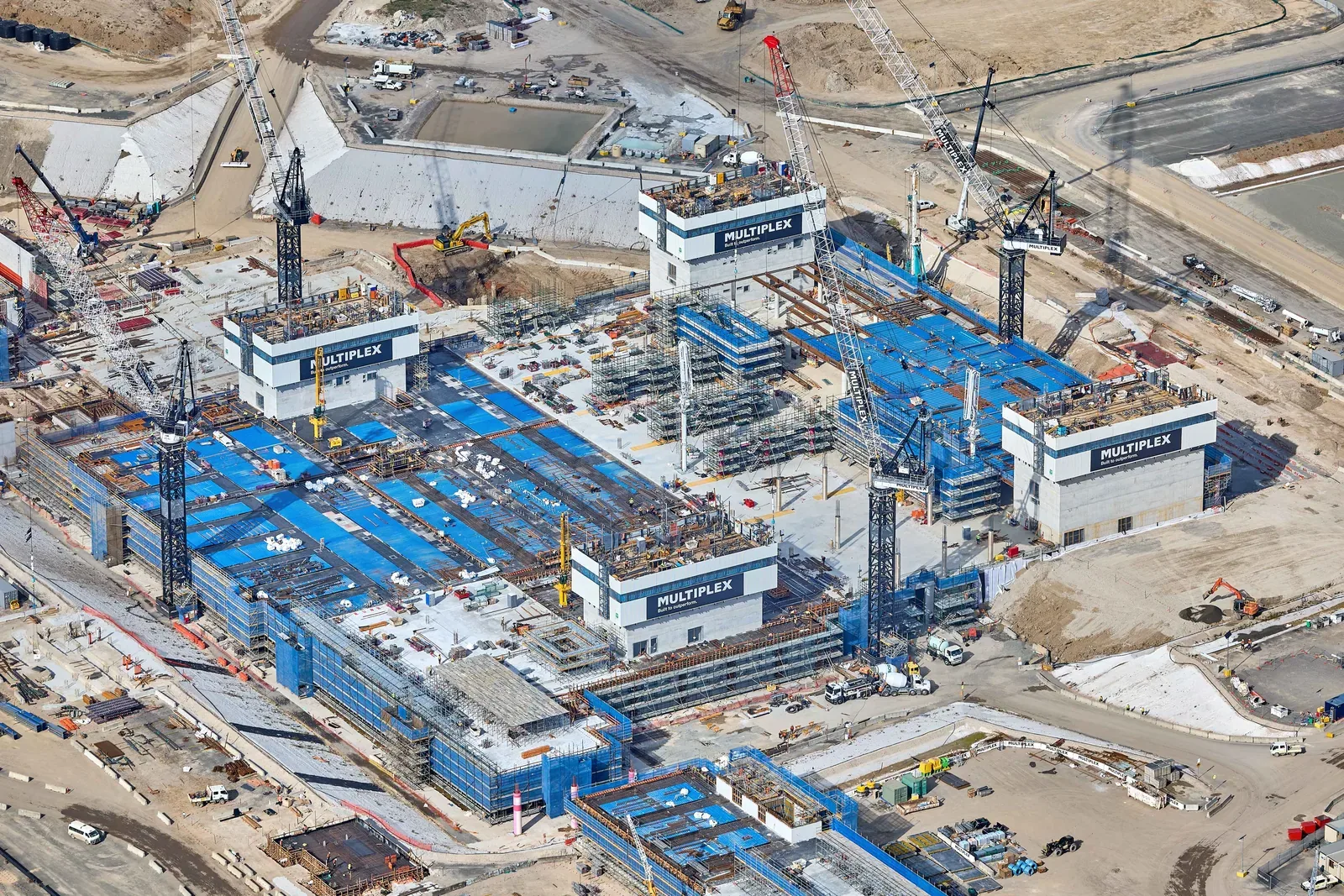 Aerial view of a large airport terminal under construction, with tower cranes, scaffolding and blue-sheeted building sections spread across the site