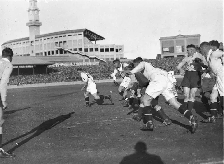 Black-and-white archive photo of rugby league players in white jerseys charging forward during a Test match, with a packed grandstand and clock tower behind