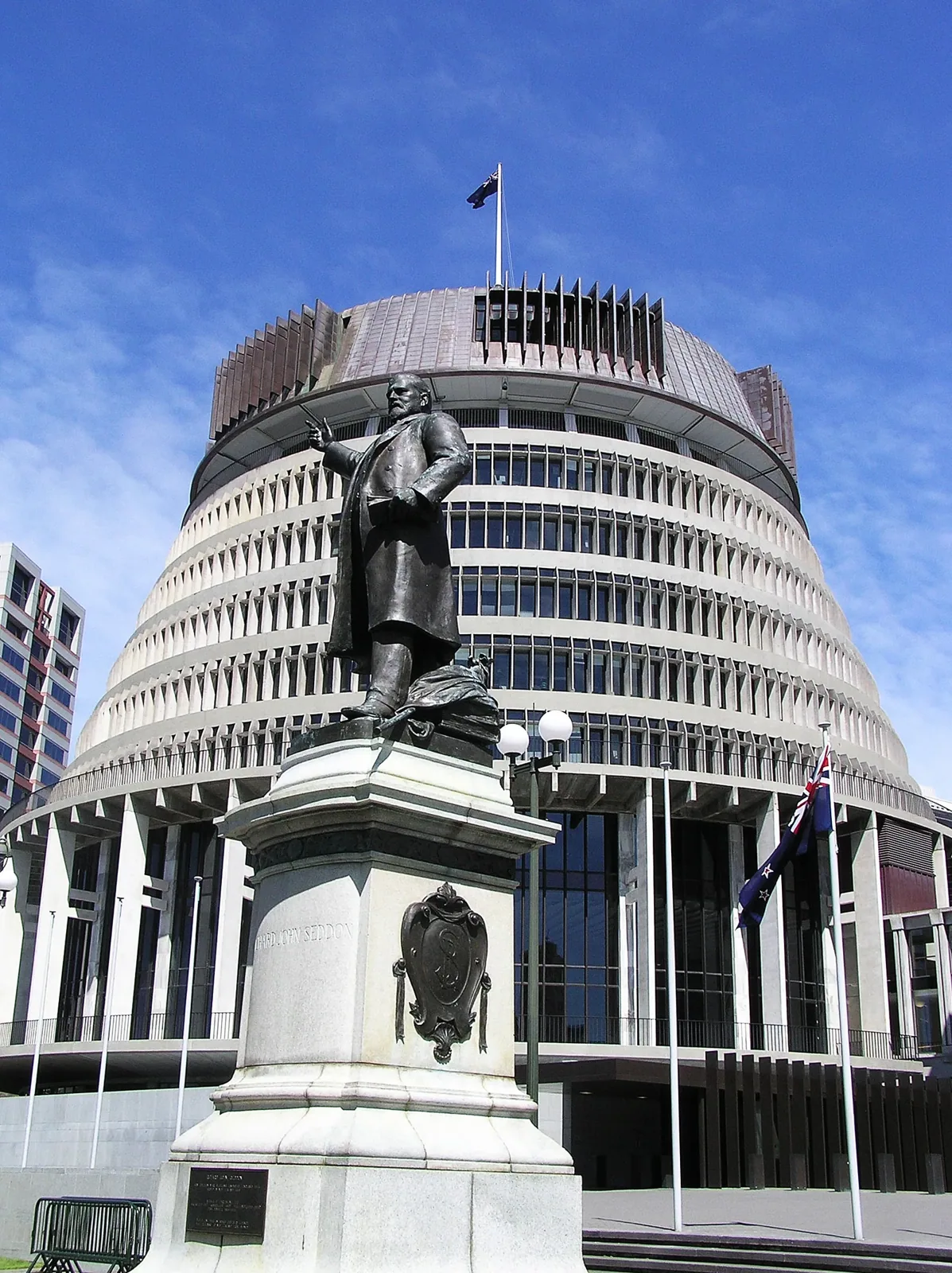 The circular, tiered Beehive building in Wellington under a blue sky, with a bronze statue of Richard John Seddon on a stone plinth in the foreground and a New Zealand flag flying beside the building