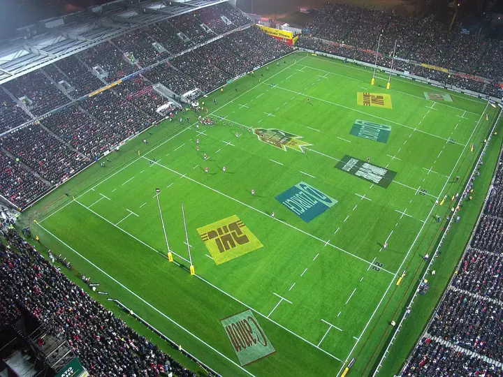 High aerial view of a floodlit rugby stadium packed with spectators, with a match under way on a green pitch marked with sponsor logos
