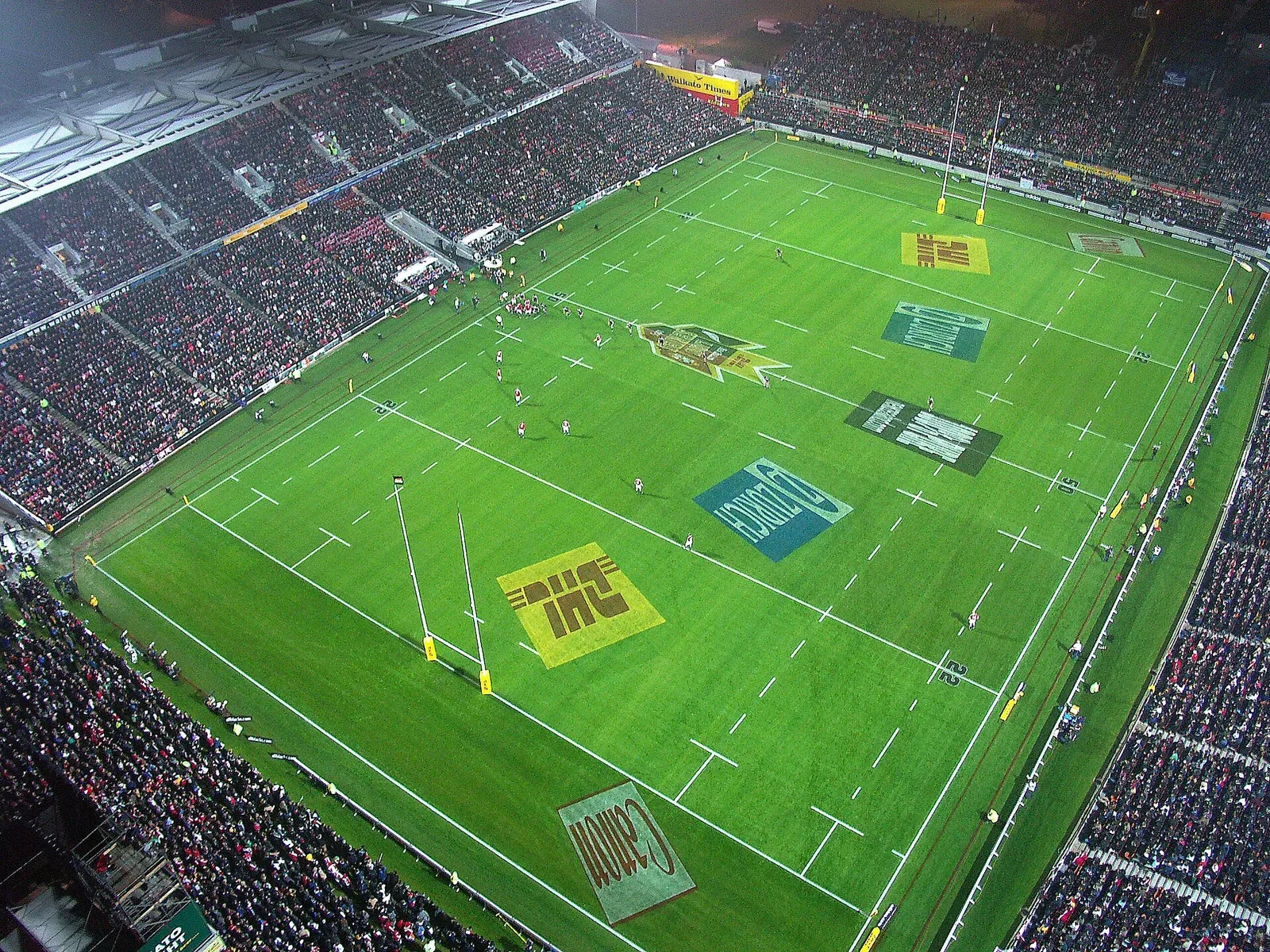 High aerial view of a floodlit rugby stadium packed with spectators, with a match under way on a green pitch marked with sponsor logos