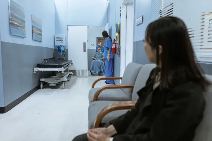 A woman in a dark coat sits on a waiting-room chair in a hospital corridor, while further down a staff member in blue scrubs stands beside a patient in a wheelchair, near an empty hospital trolley