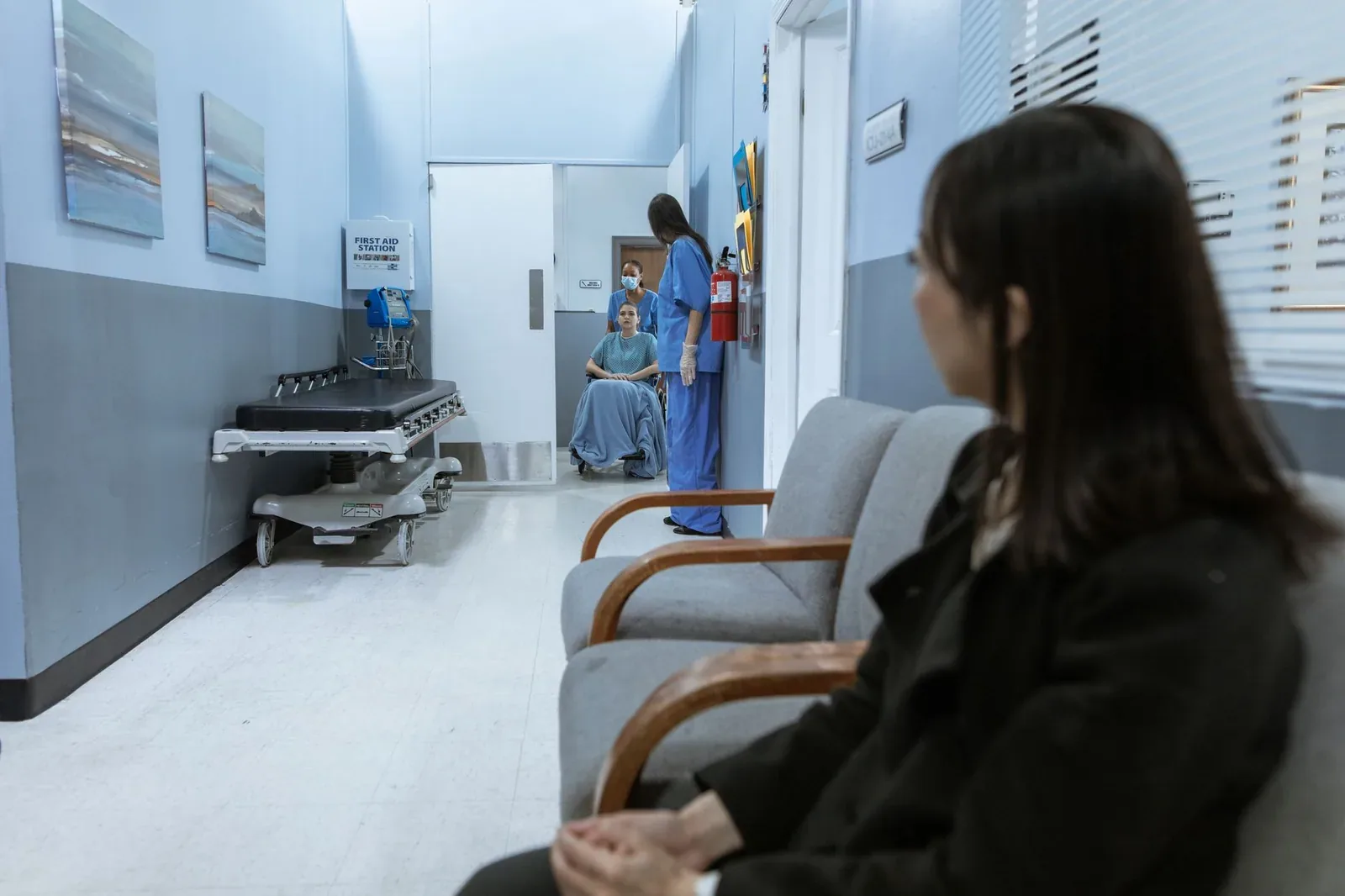 A woman in a dark coat sits on a waiting-room chair in a hospital corridor, while further down a staff member in blue scrubs stands beside a patient in a wheelchair, near an empty hospital trolley