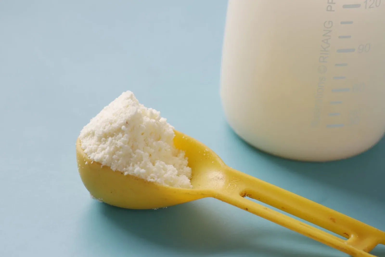 A yellow measuring scoop heaped with white milk powder beside a plastic measuring bottle on a pale blue background
