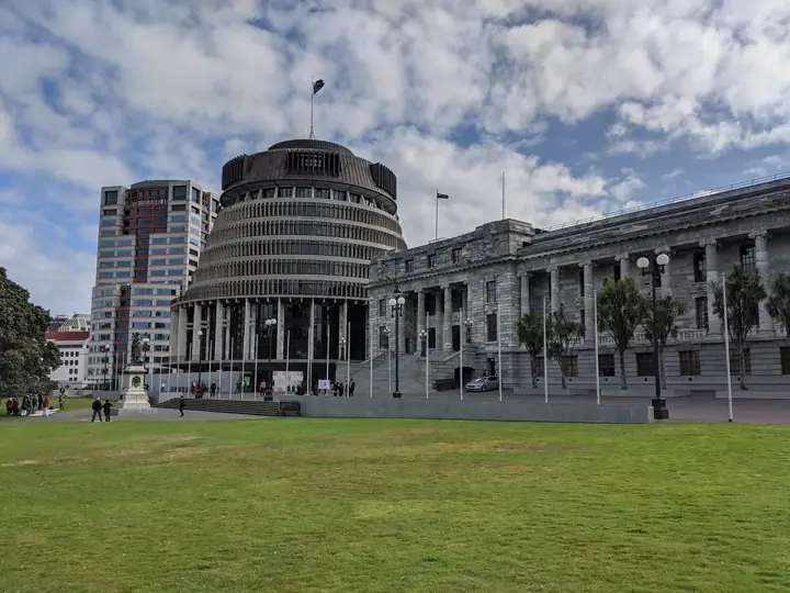 The round Beehive building next to the stone Parliament House in Wellington, seen across a wide lawn under a cloudy blue sky