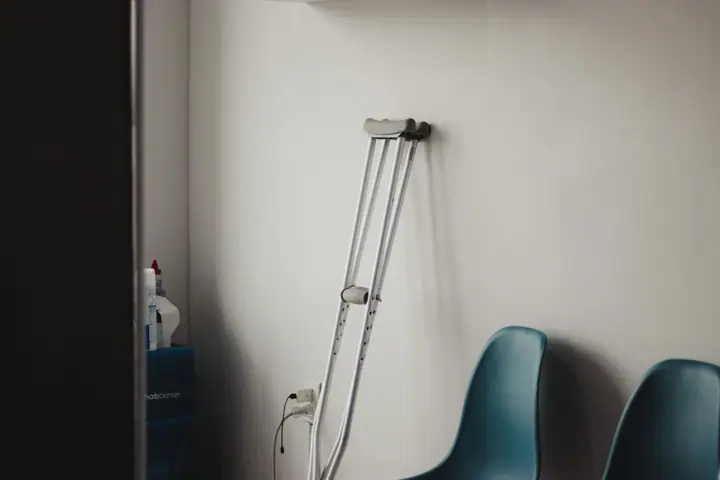 A pair of crutches leaning against a plain white wall beside two empty blue plastic chairs in a sparse waiting room