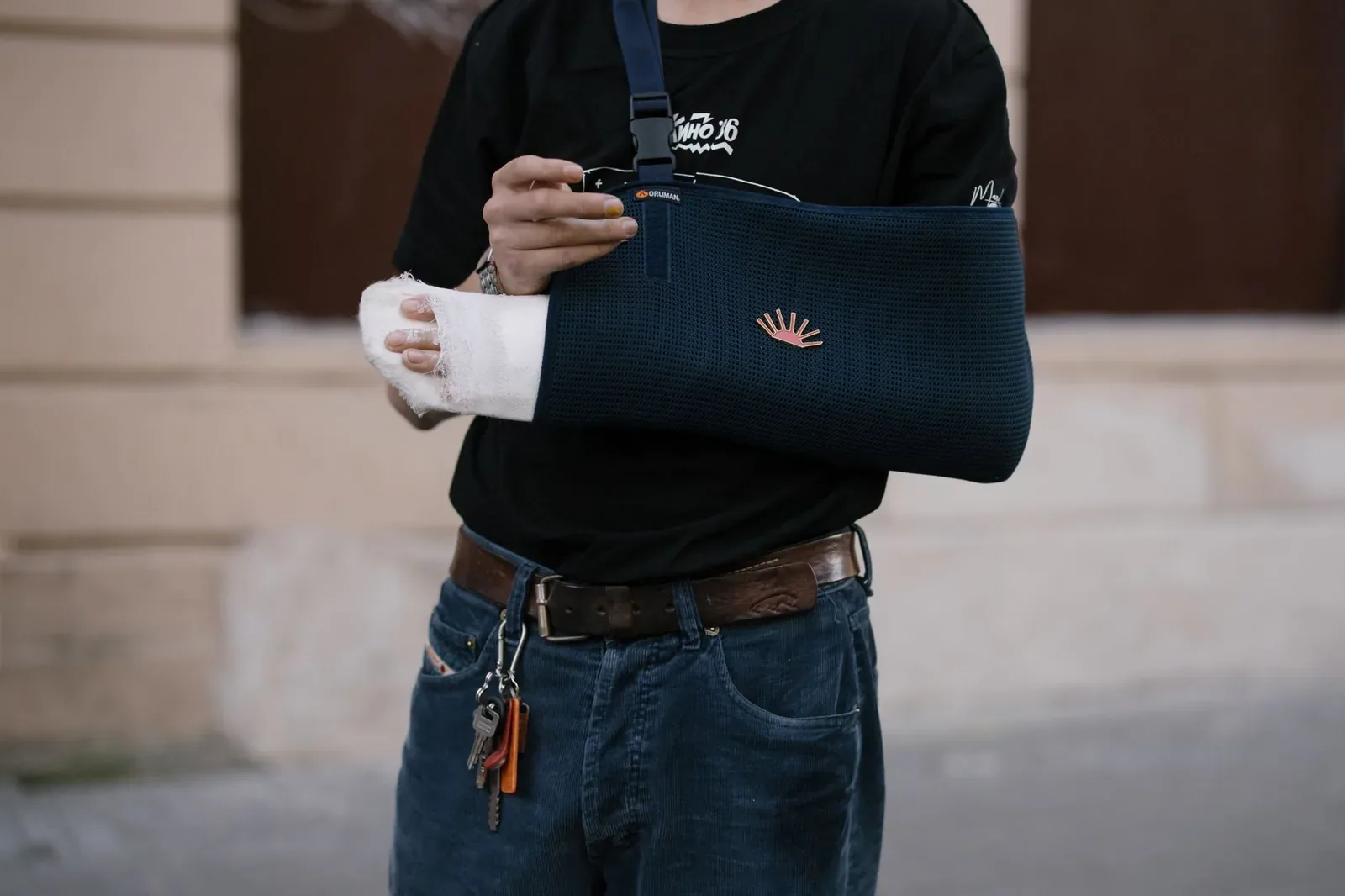 A man in a black T-shirt and jeans, face out of frame, with his bandaged forearm supported in a dark arm sling