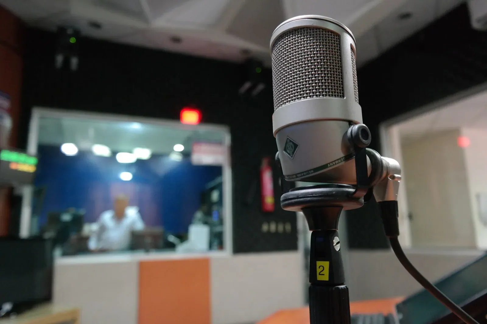 Close-up of a condenser microphone on a stand in a broadcast studio, with a blurred engineer behind a glass window in the background