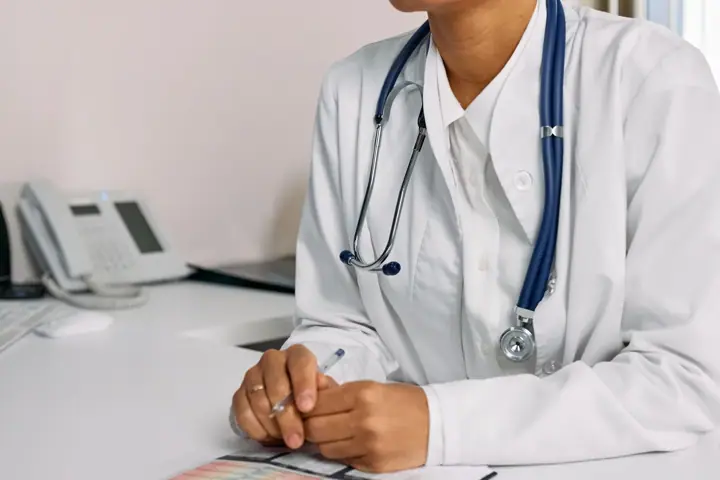 A doctor in a white coat with a stethoscope round their neck sits at a clinic desk holding a pen, with a desk phone beside them. The face is out of frame.