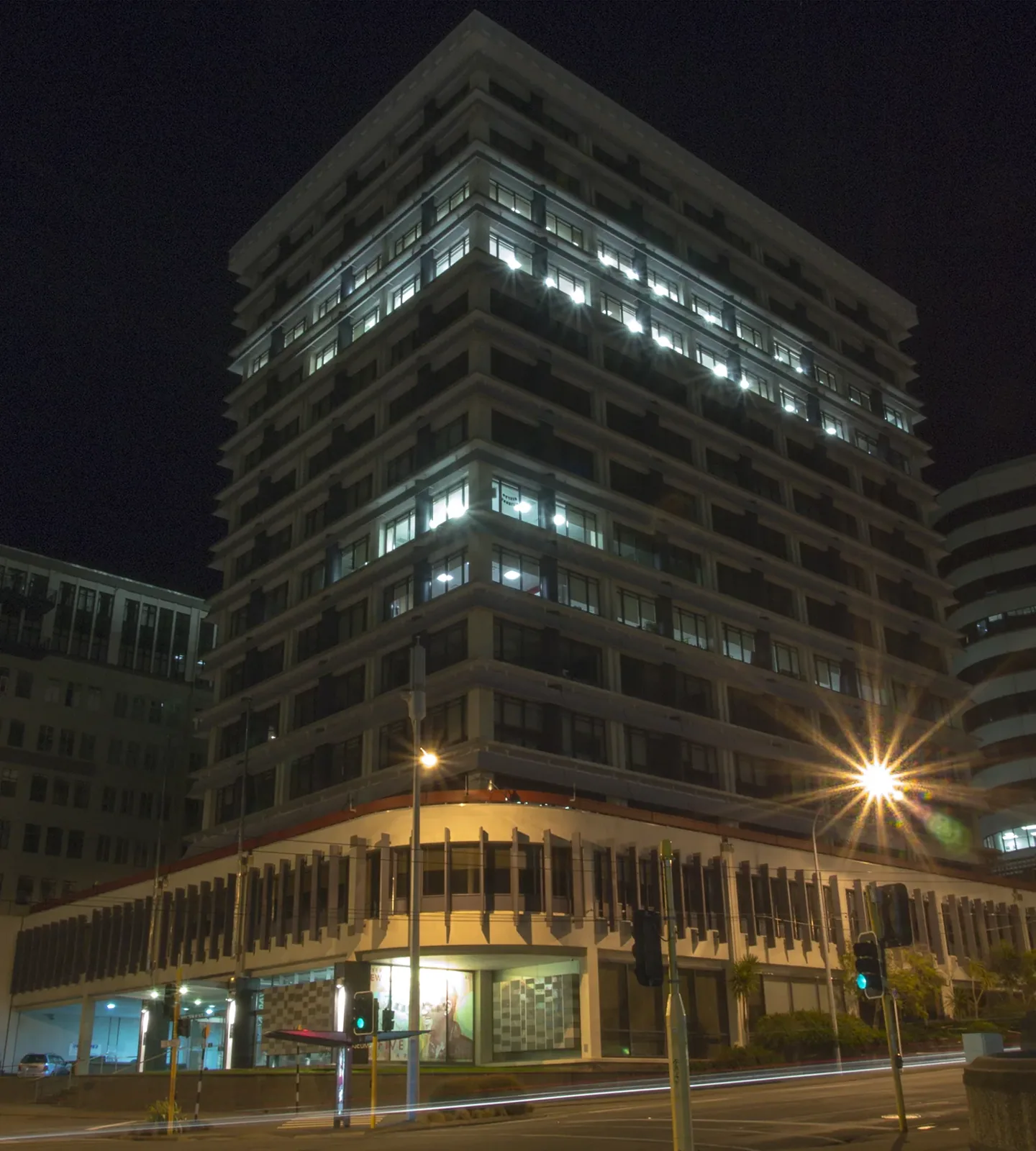 A multi-storey office tower lit up at night above a low colonnaded podium, with street lights and traffic signals in the foreground
