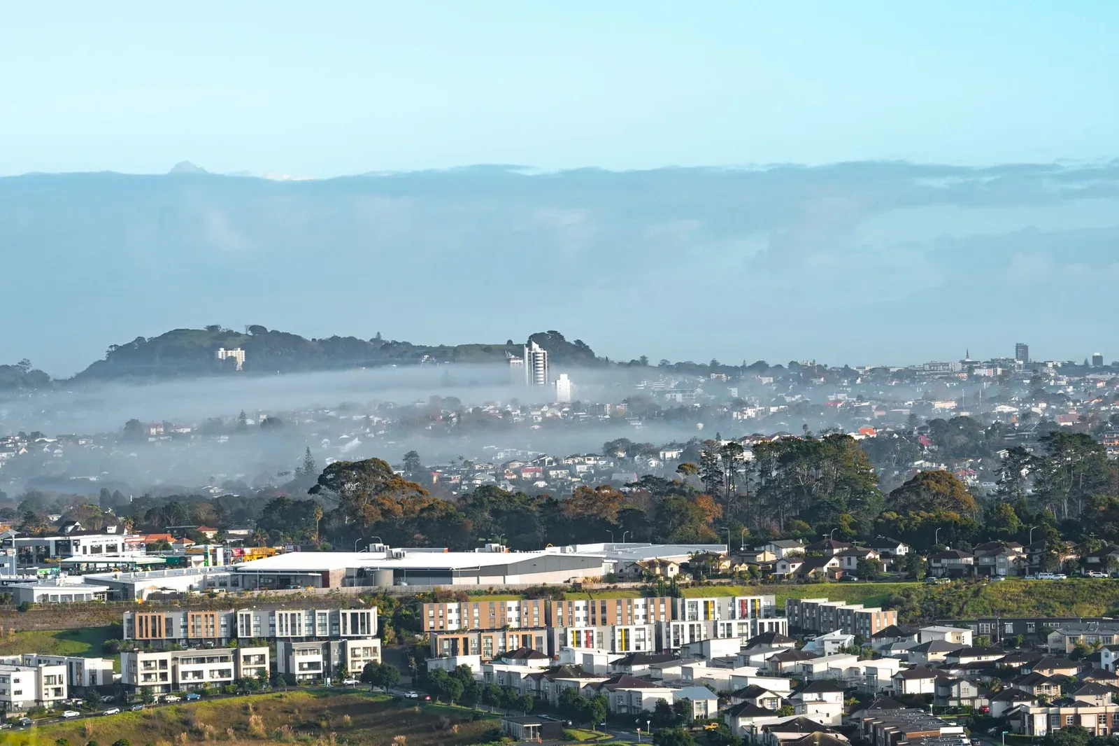Elevated view over rows of new townhouses and suburban homes, with morning fog lying over the city and a volcanic hill behind
