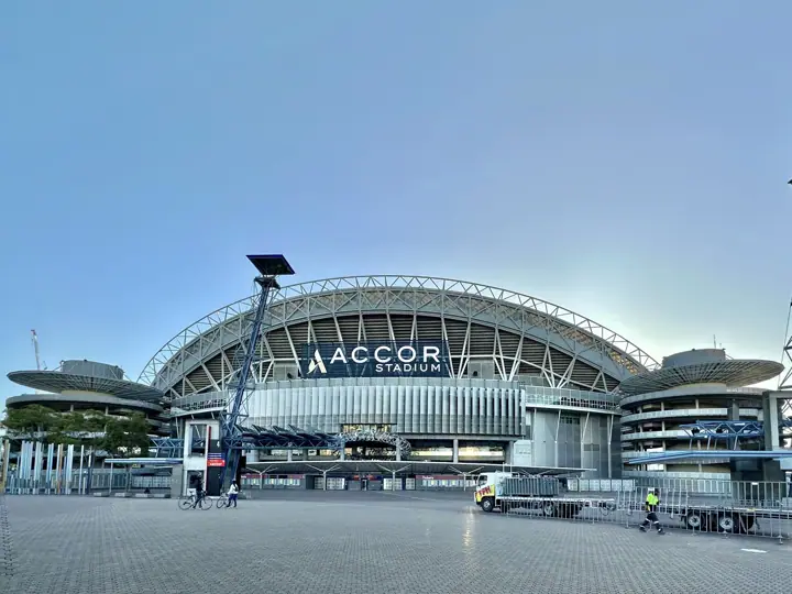Exterior of Accor Stadium in daylight, its curved steel roof above the 'Accor Stadium' sign, with an empty paved forecourt in front