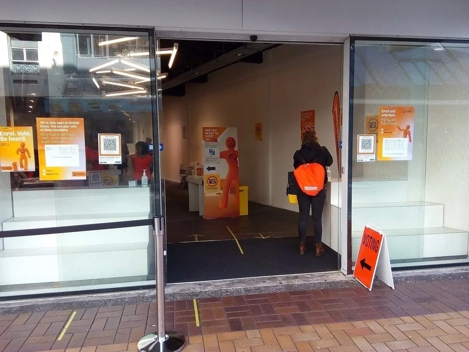 Entrance to a shopfront advance voting place with orange 'Enrol. Vote. Be heard' posters and an orange 'Voting' sign on the pavement; a voter with an orange backpack stands inside, seen from behind.
