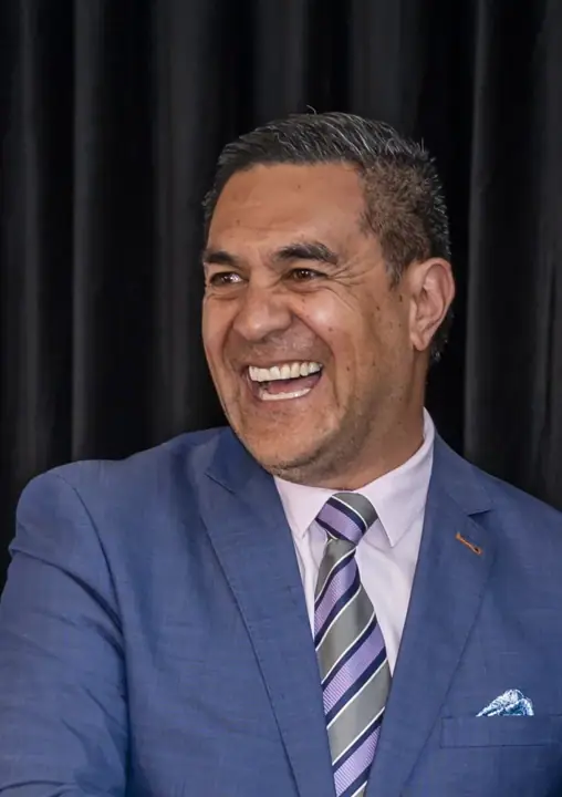A smiling man in a blue suit, light pink shirt and striped purple tie, against a dark curtain