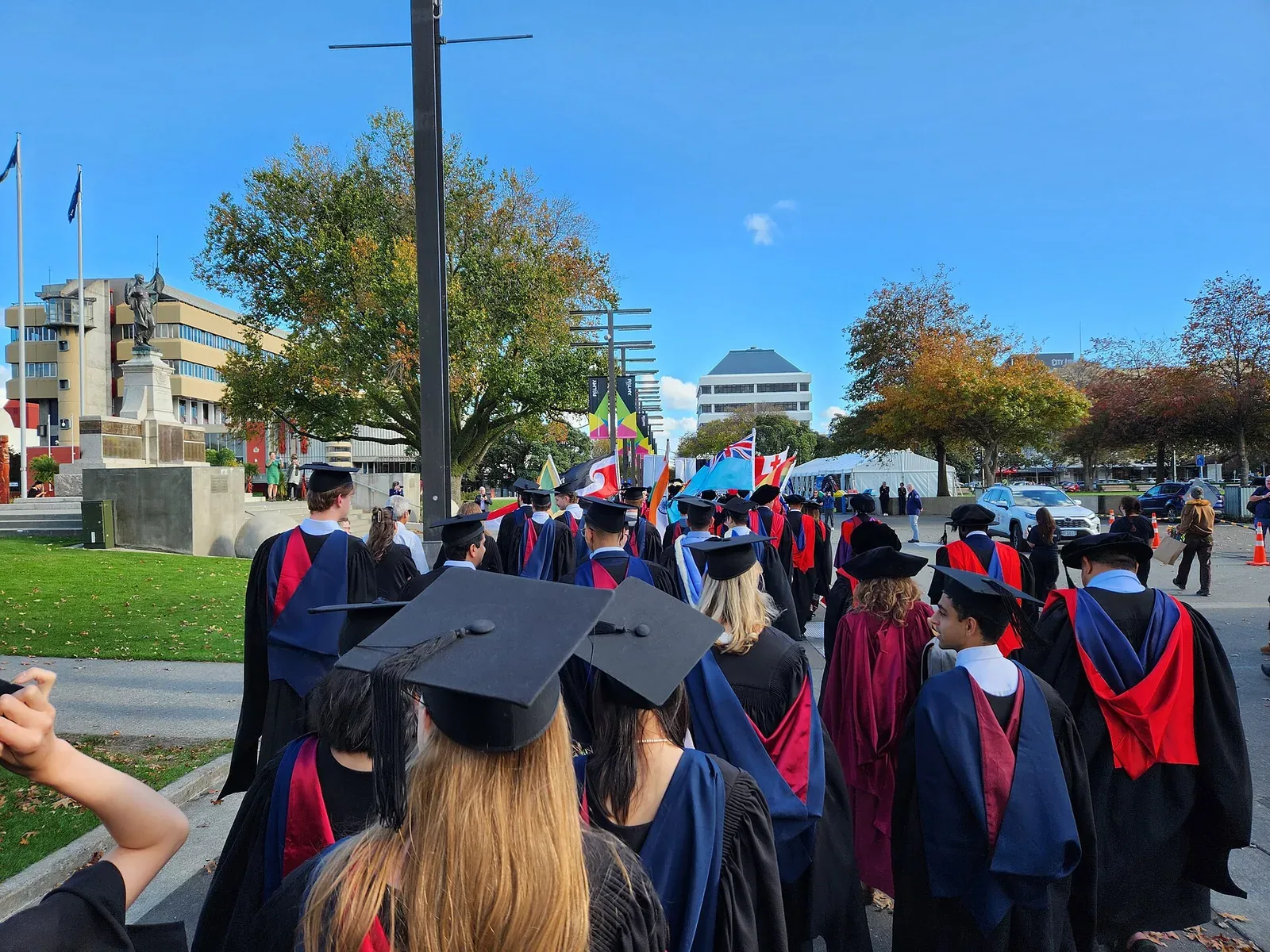 Graduates in black gowns, mortarboards and coloured hoods walk away from the camera in a procession along a city street on a sunny day, with trees and buildings ahead.