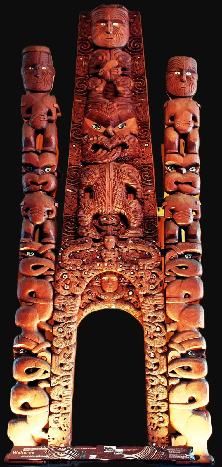 A tall, ornately carved red-brown Māori entrance gateway with stacked human figures and a central arched opening, shown against a black background