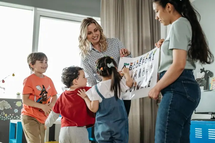 Two women hold up a hand-drawn alphabet chart while four young children gather round in a bright classroom