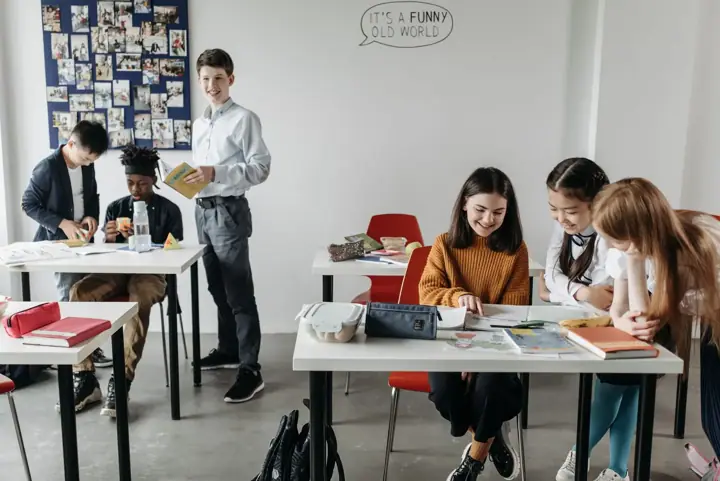 Secondary-age students sit at white desks in a bright classroom, smiling over books and papers, while another student stands holding a book