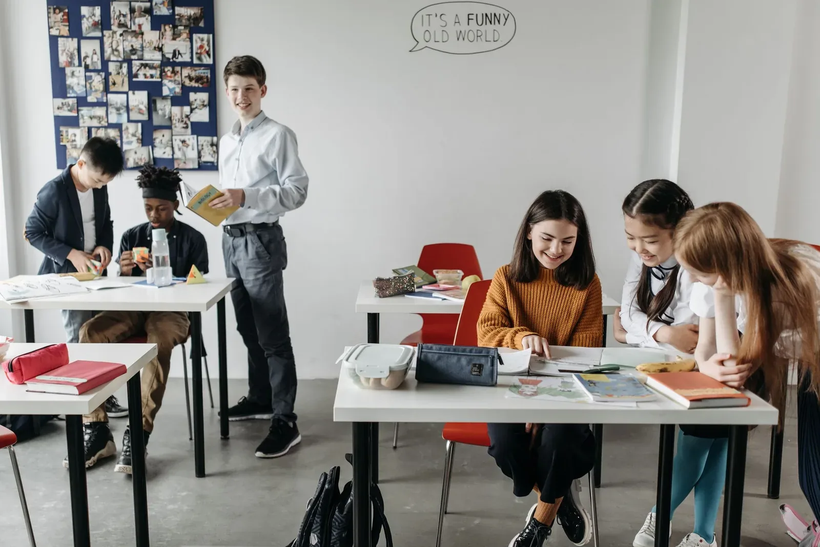 Secondary-age students sit at white desks in a bright classroom, smiling over books and papers, while another student stands holding a book