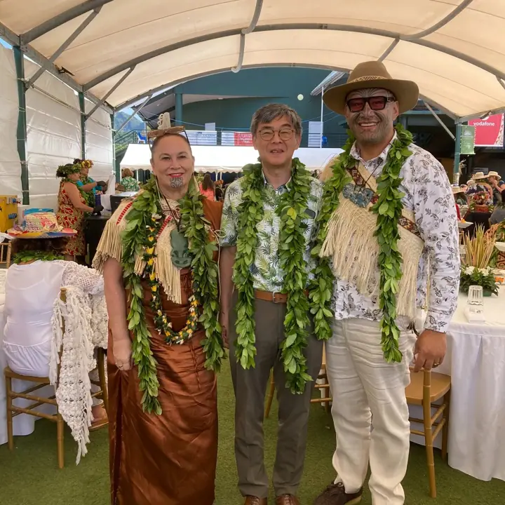 Three people stand together under a white marquee wearing green leaf garlands; on the right a man in a wide-brimmed hat, sunglasses and a fringed cloak.