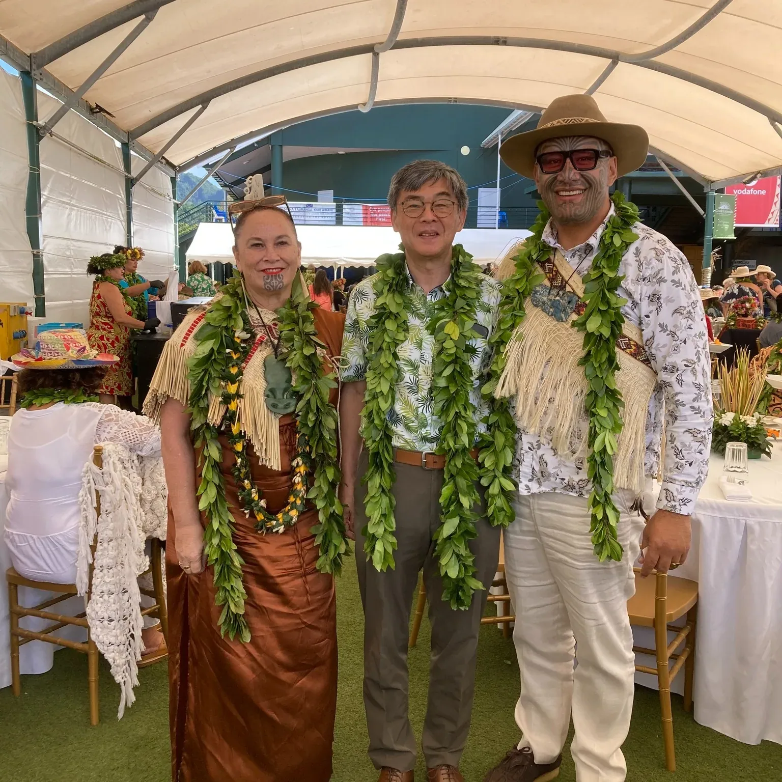 Three people stand together under a white marquee wearing green leaf garlands; on the right a man in a wide-brimmed hat, sunglasses and a fringed cloak.