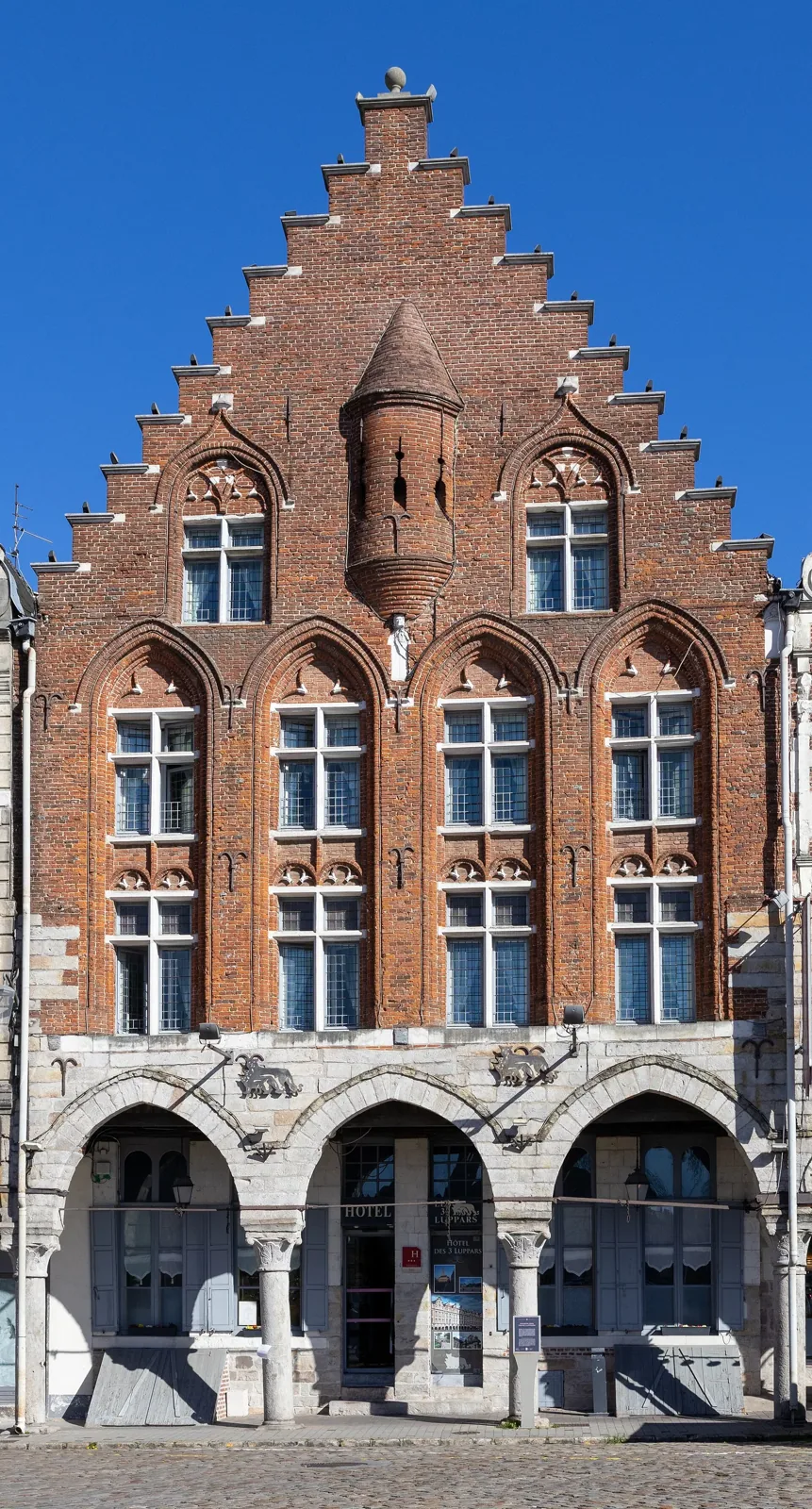 The tall red-brick stepped gable of the Maison des Trois Luppars, with a small turret and stone arcades at street level, under a clear blue sky