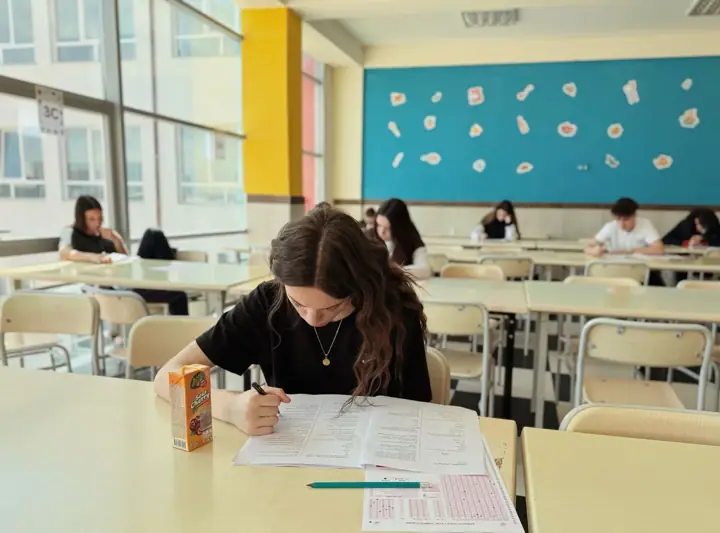 A student with long dark hair, head bowed, fills in an exam answer sheet at a desk in a bright classroom while other students work at tables behind her